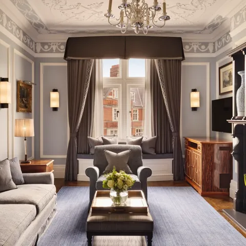 Elegant living room with grey sofa and armchairs, fireplace, window seat with cushions, and large window with drapes.