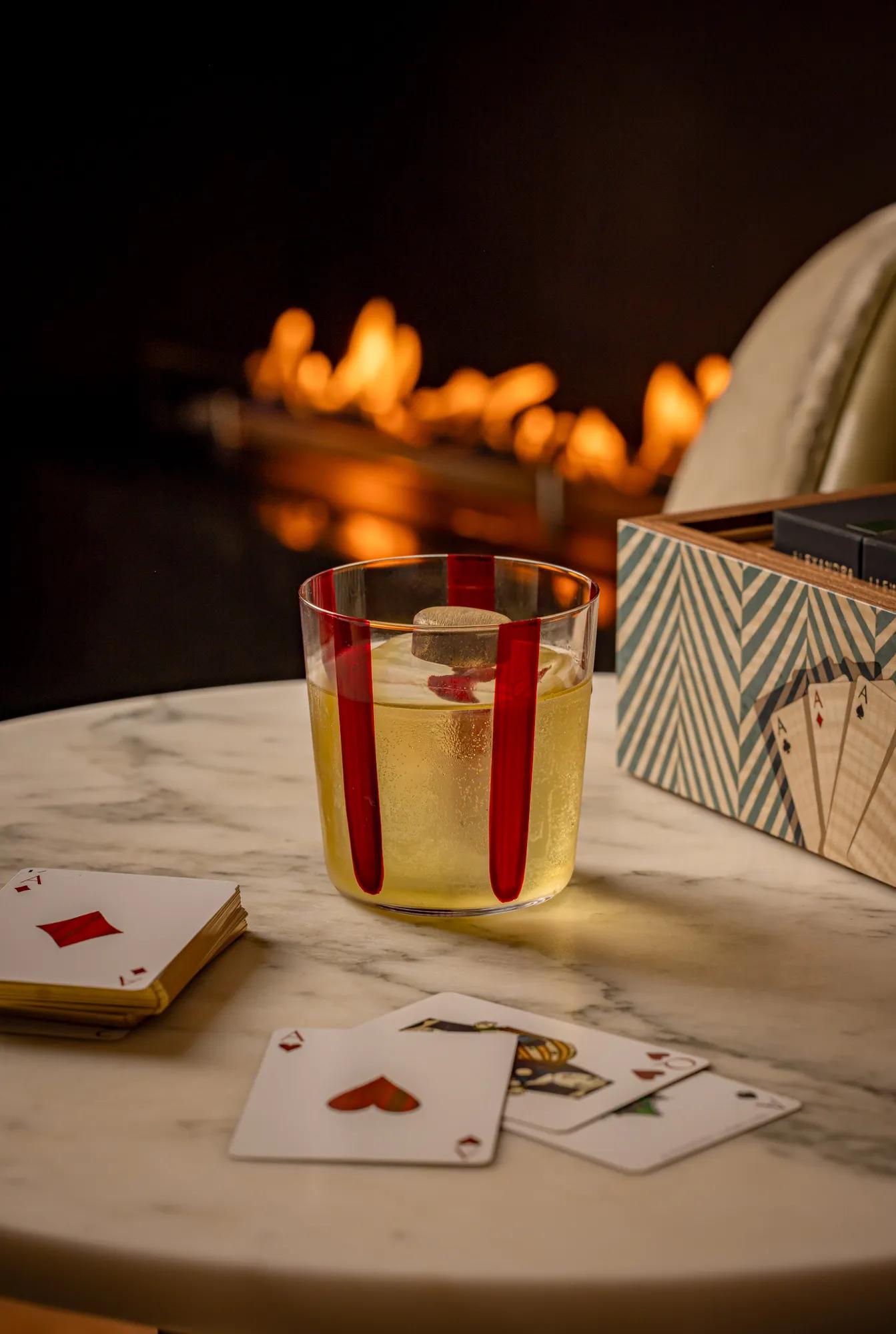 Glass of sparkling drink beside playing cards on a marble table, with a softly glowing fireplace blurred in the background.