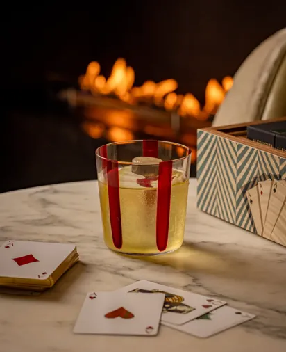 Glass of sparkling drink beside playing cards on a marble table, with a softly glowing fireplace blurred in the background.