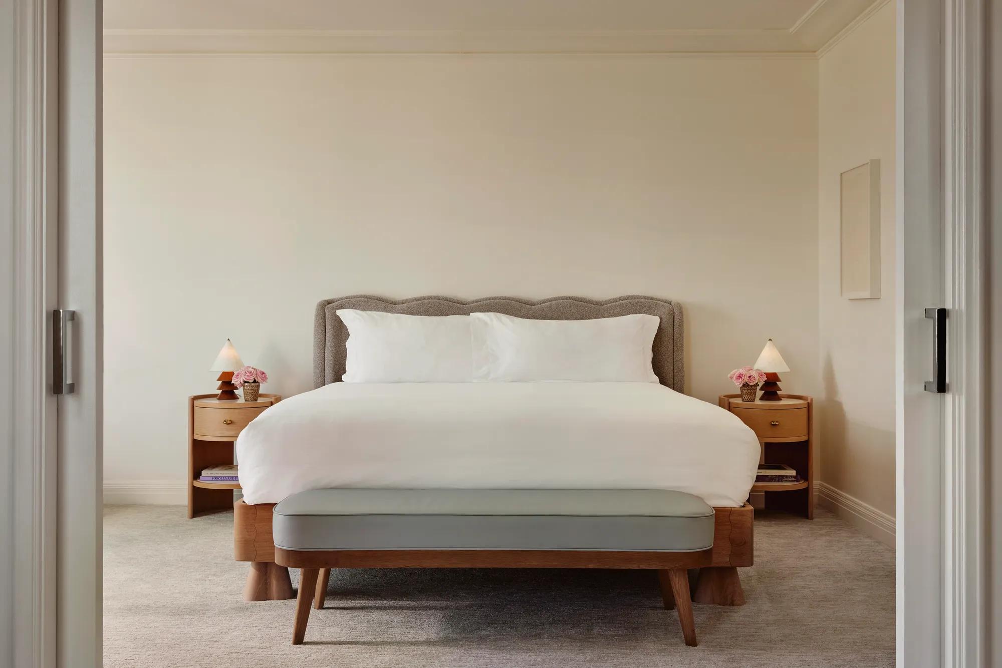 Bright Maybourne Beverly Hills Garden  suite bedroom with plush king bed, grey headboard, bench, twin nightstands, lamps, and pink roses.