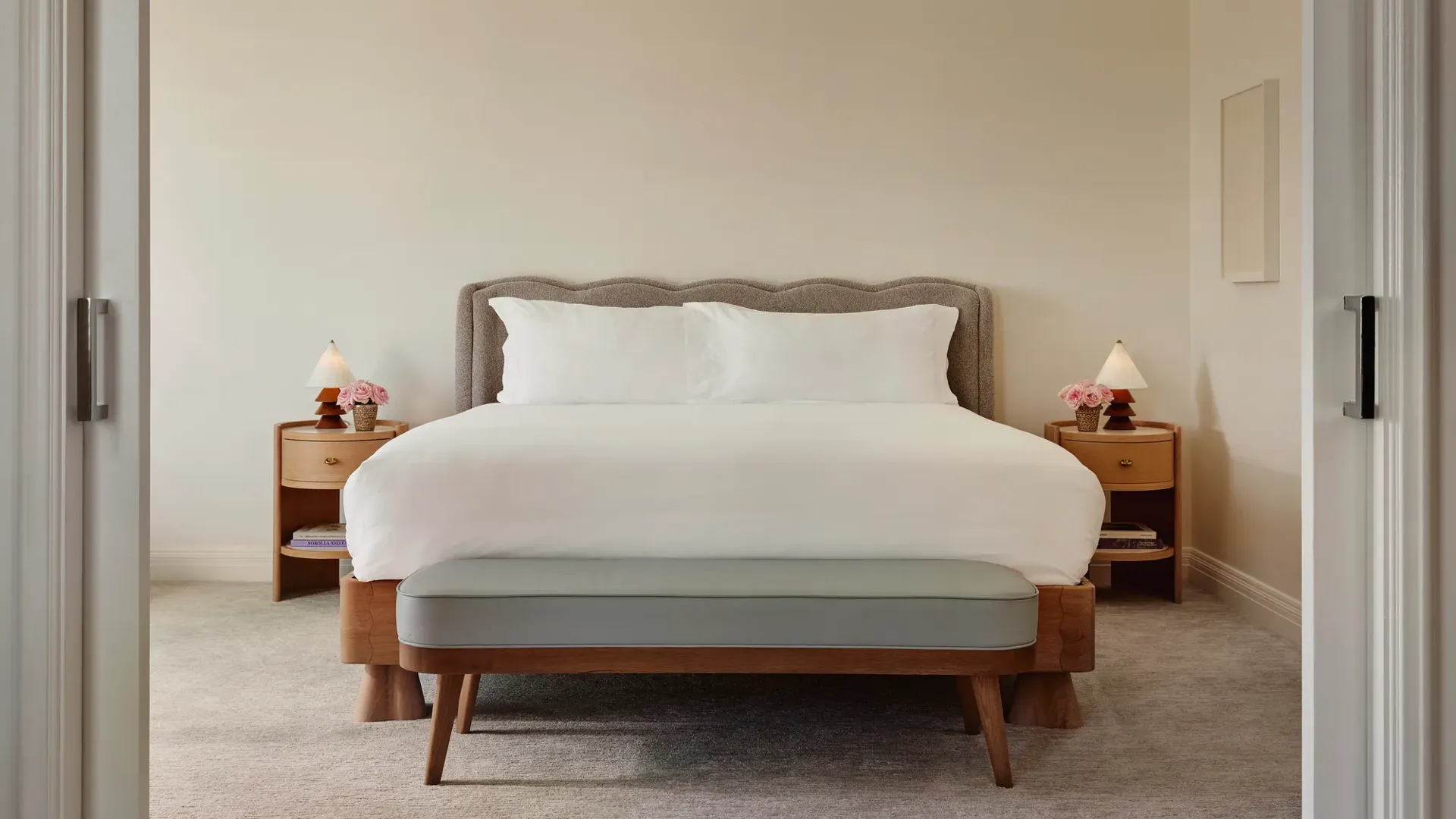 Bright Maybourne Beverly Hills Garden  suite bedroom with plush king bed, grey headboard, bench, twin nightstands, lamps, and pink roses.