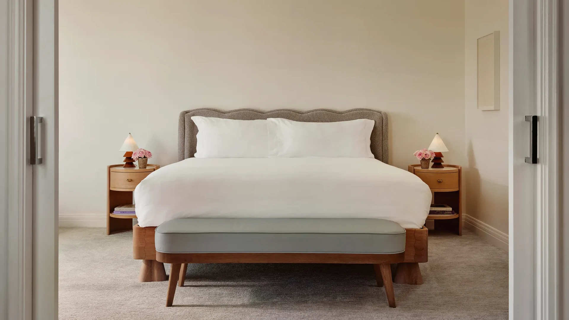 Bright Maybourne Beverly Hills Garden  suite bedroom with plush king bed, grey headboard, bench, twin nightstands, lamps, and pink roses.