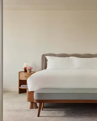 Bright Maybourne Beverly Hills Garden  suite bedroom with plush king bed, grey headboard, bench, twin nightstands, lamps, and pink roses.