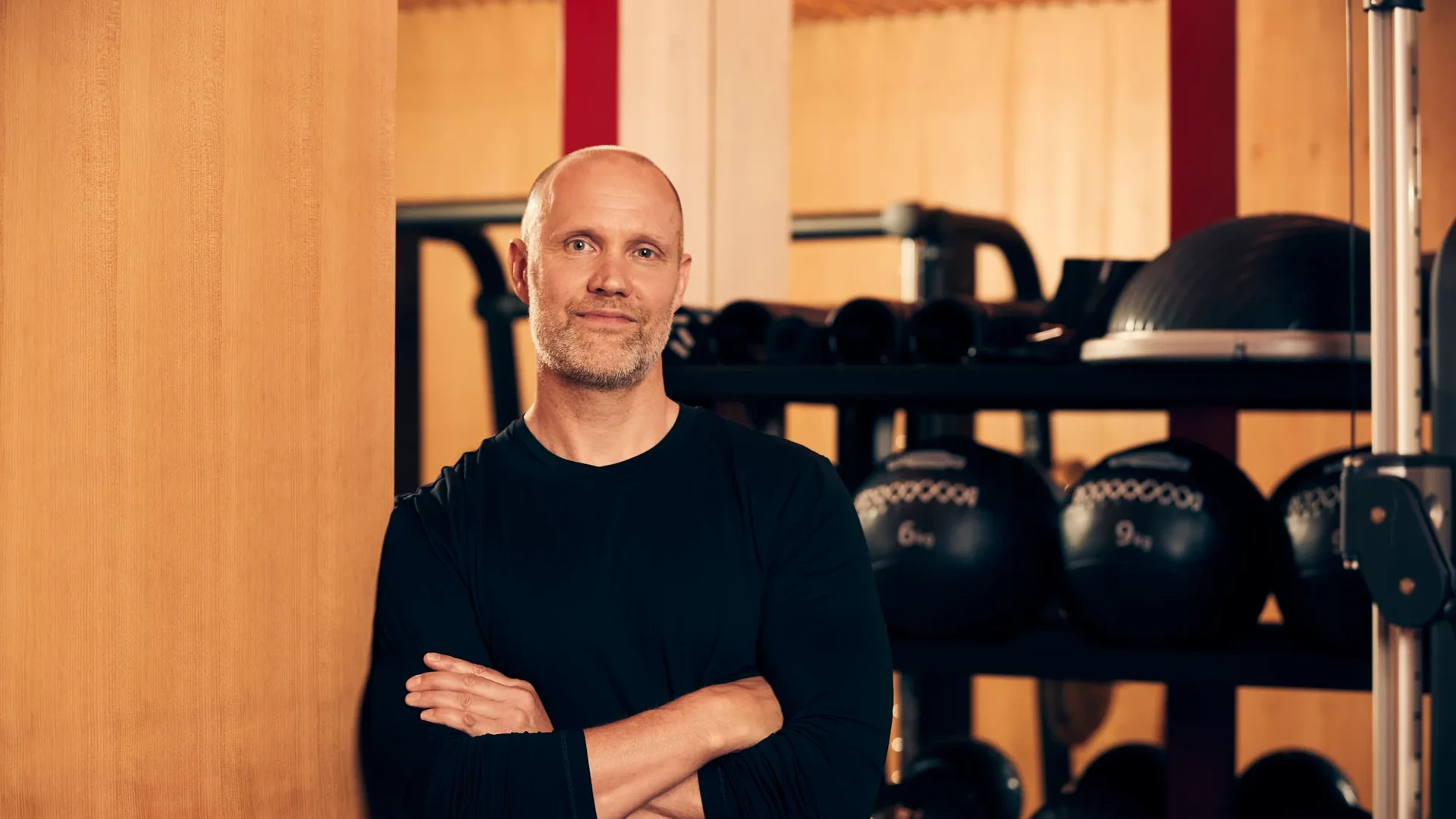 Male fitness instructor standing with arms crossed in a gym, with medicine balls and training equipment behind him.