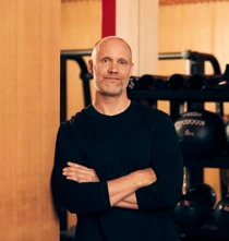Male fitness instructor standing with arms crossed in a gym, with medicine balls and training equipment behind him.