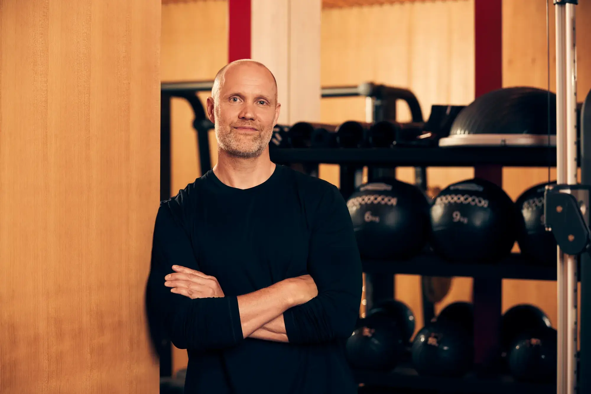 Male fitness instructor standing with arms crossed in a gym, with medicine balls and training equipment behind him.
