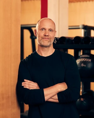 Male fitness instructor standing with arms crossed in a gym, with medicine balls and training equipment behind him.