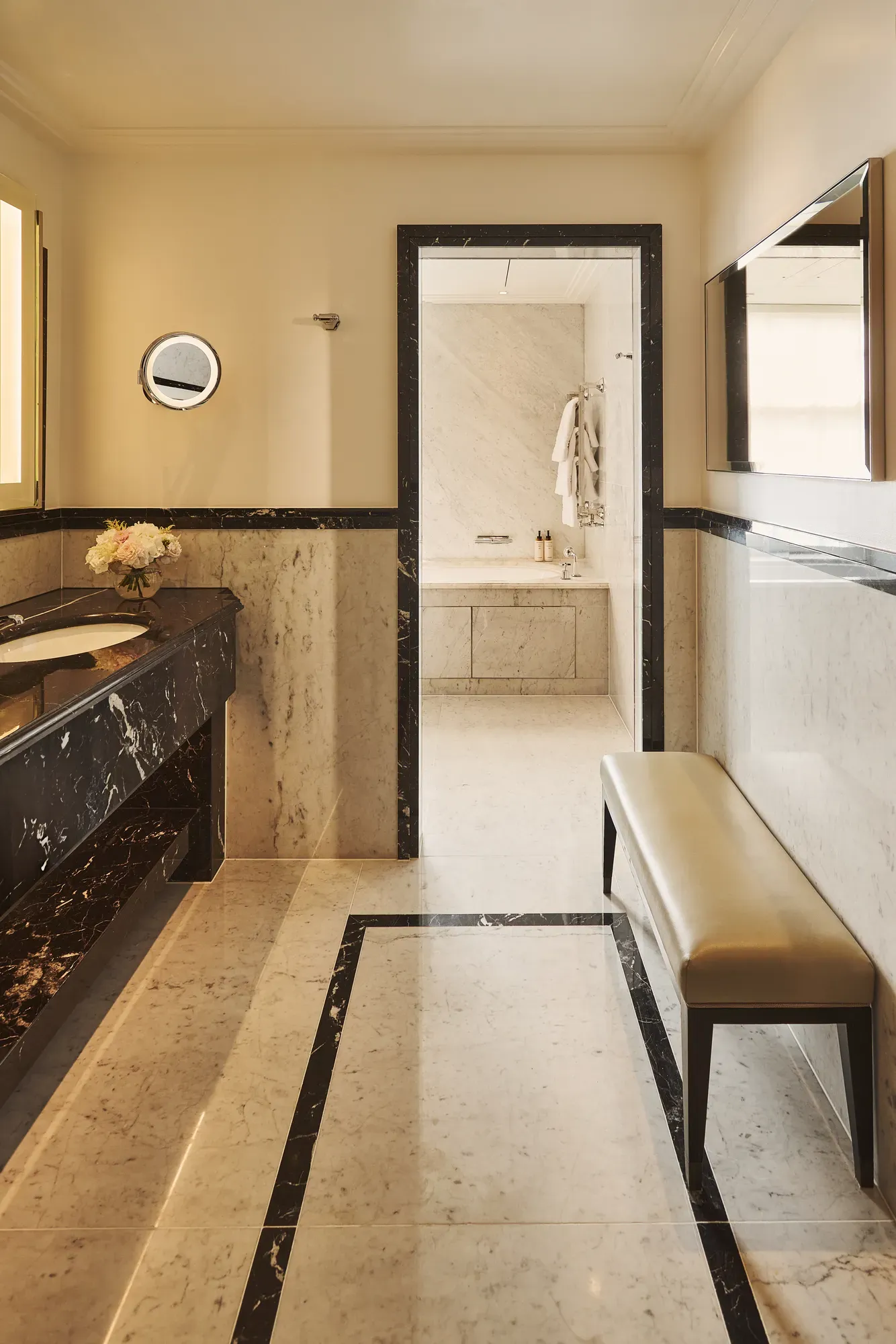 Marble bathroom with double vanity, mirror and bench, leading to walk-in shower area, finished in neutral tones with soft lighting.
