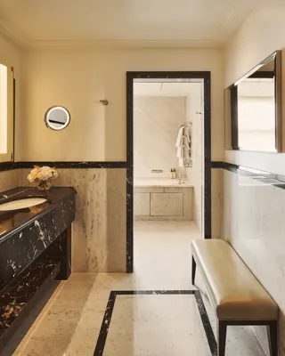 Marble bathroom with double vanity, mirror and bench, leading to walk-in shower area, finished in neutral tones with soft lighting.