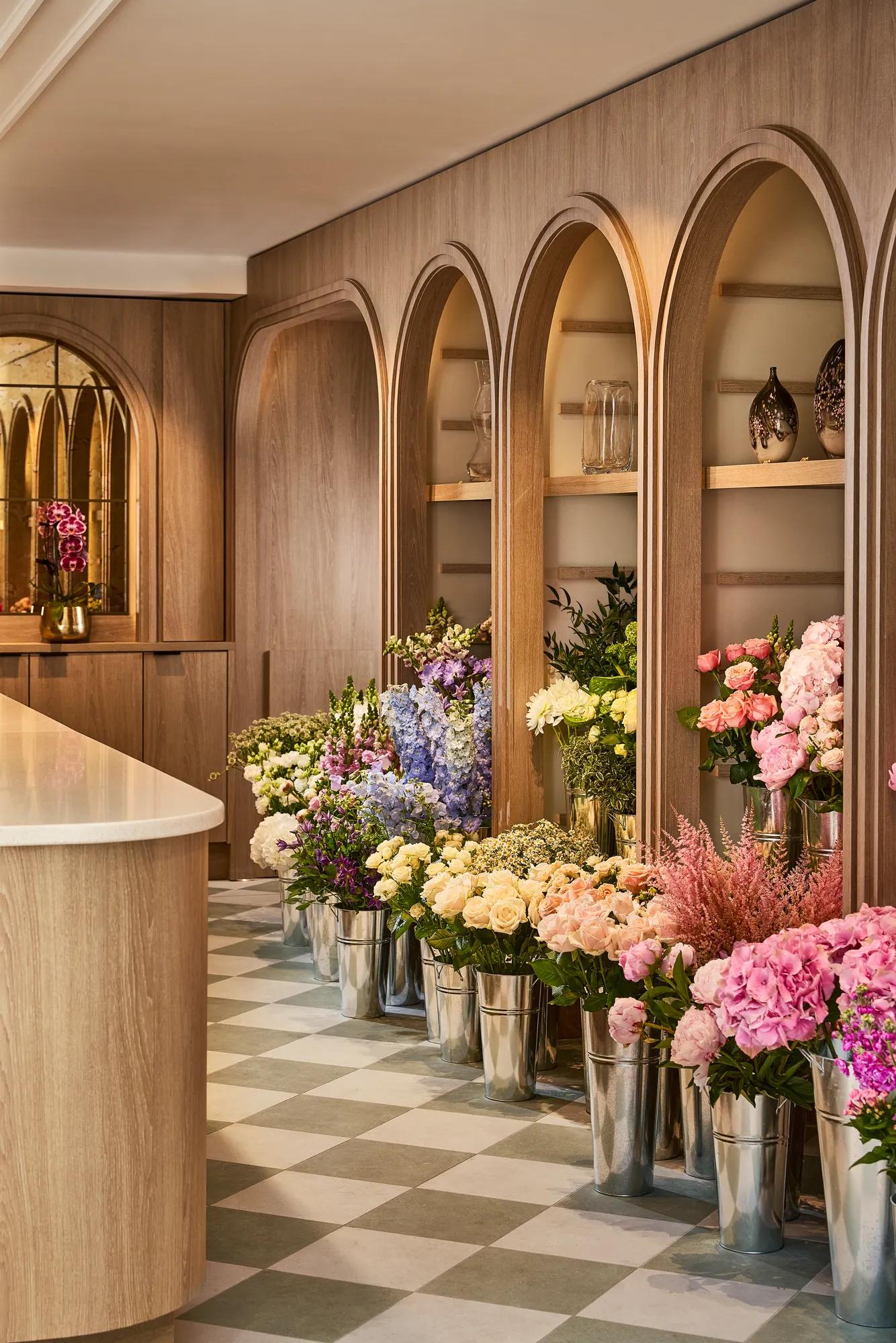 Florist boutique with rows of fresh flowers in silver buckets, set against warm oak shelving and a chequered stone floor.