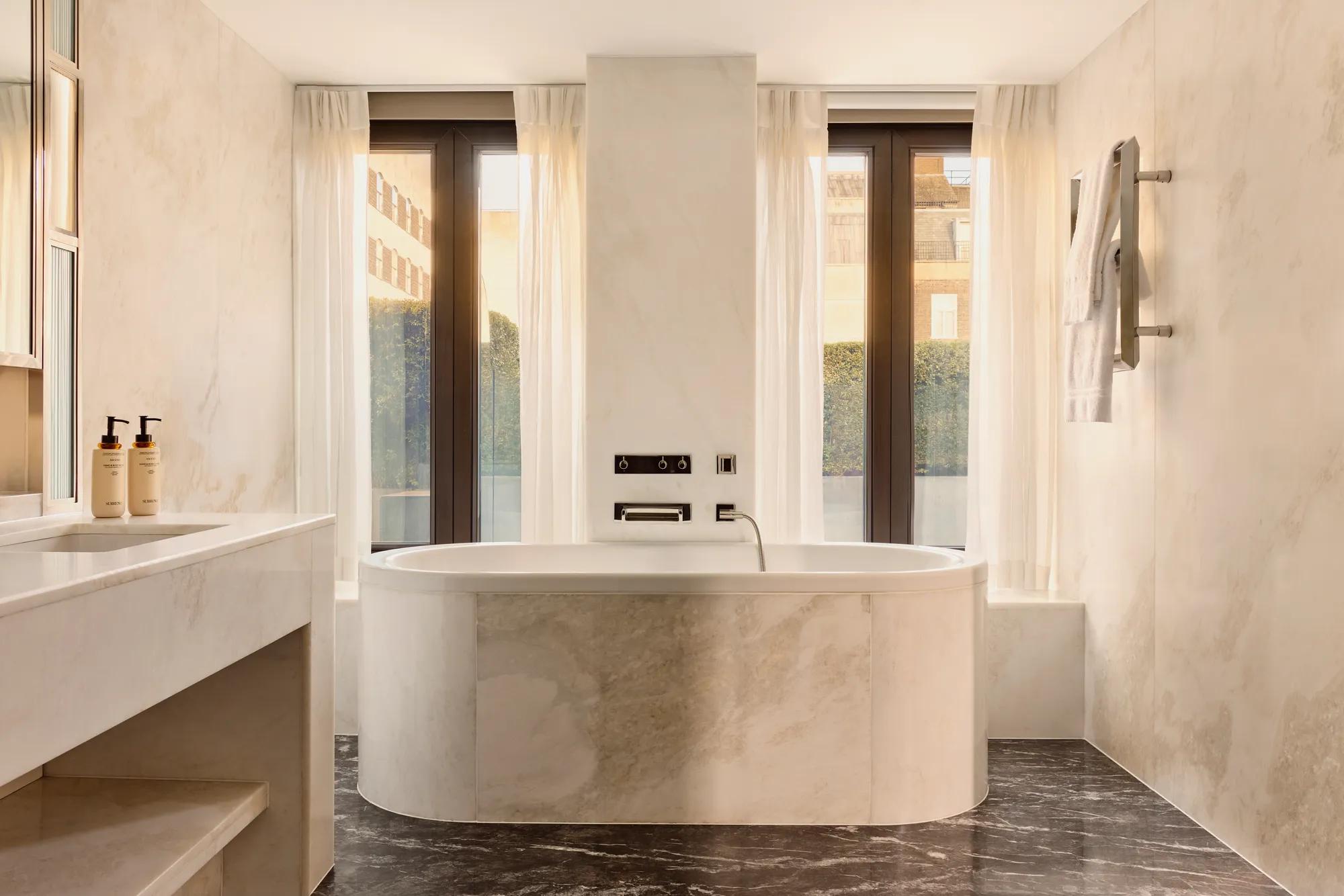 Sunlit marble bathroom with oval bathtub, large windows, towel rail, and sleek vanity area.