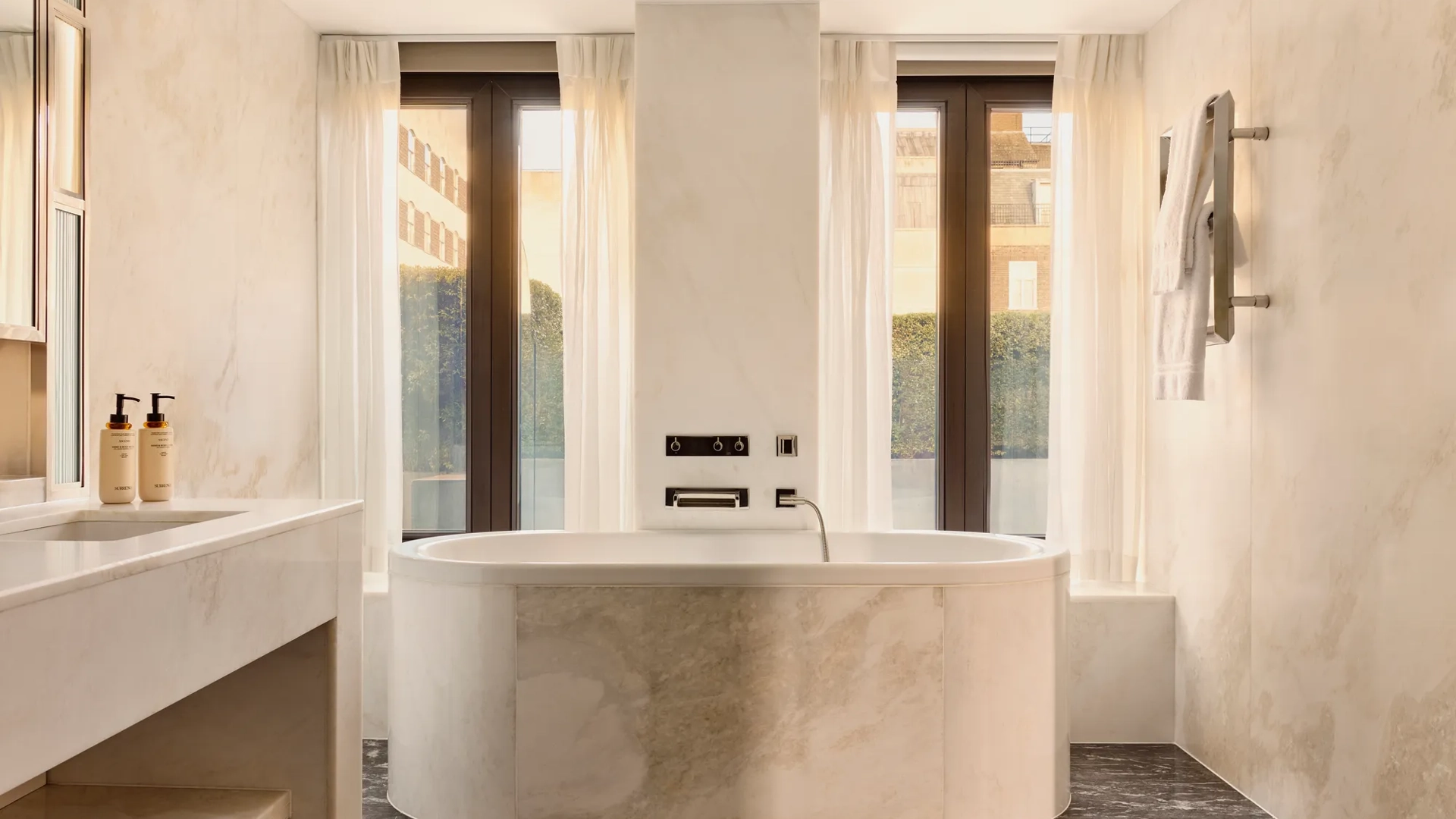 Sunlit marble bathroom with oval bathtub, large windows, towel rail, and sleek vanity area.