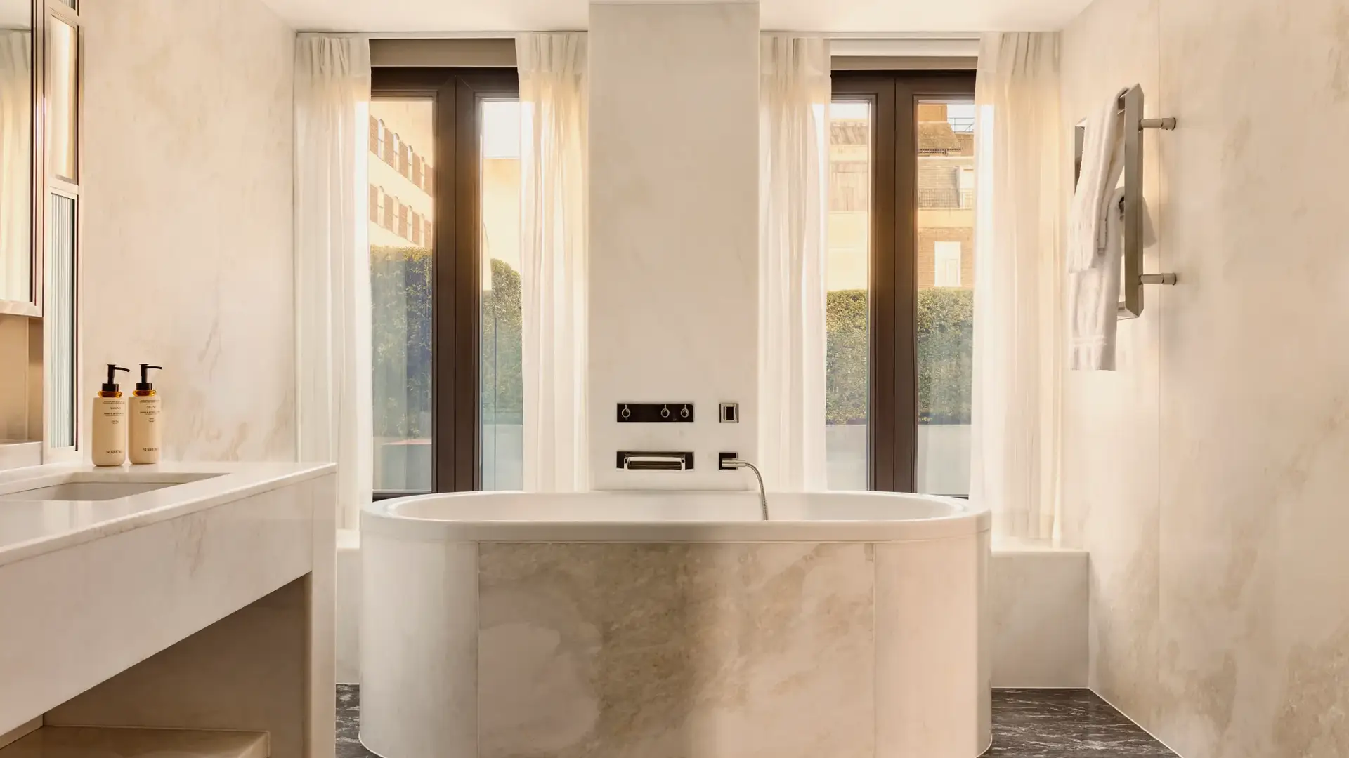 Sunlit marble bathroom with oval bathtub, large windows, towel rail, and sleek vanity area.