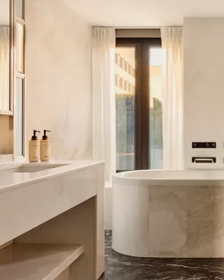 Sunlit marble bathroom with oval bathtub, large windows, towel rail, and sleek vanity area.
