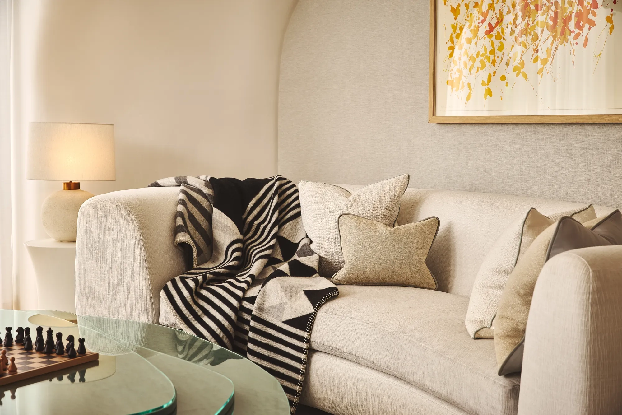 A cosy living area with a cream curved sofa layered with neutral cushions and a black-and-white geometric throw, beside a glass coffee table with a chessboard and a soft-glowing lamp on a side table.