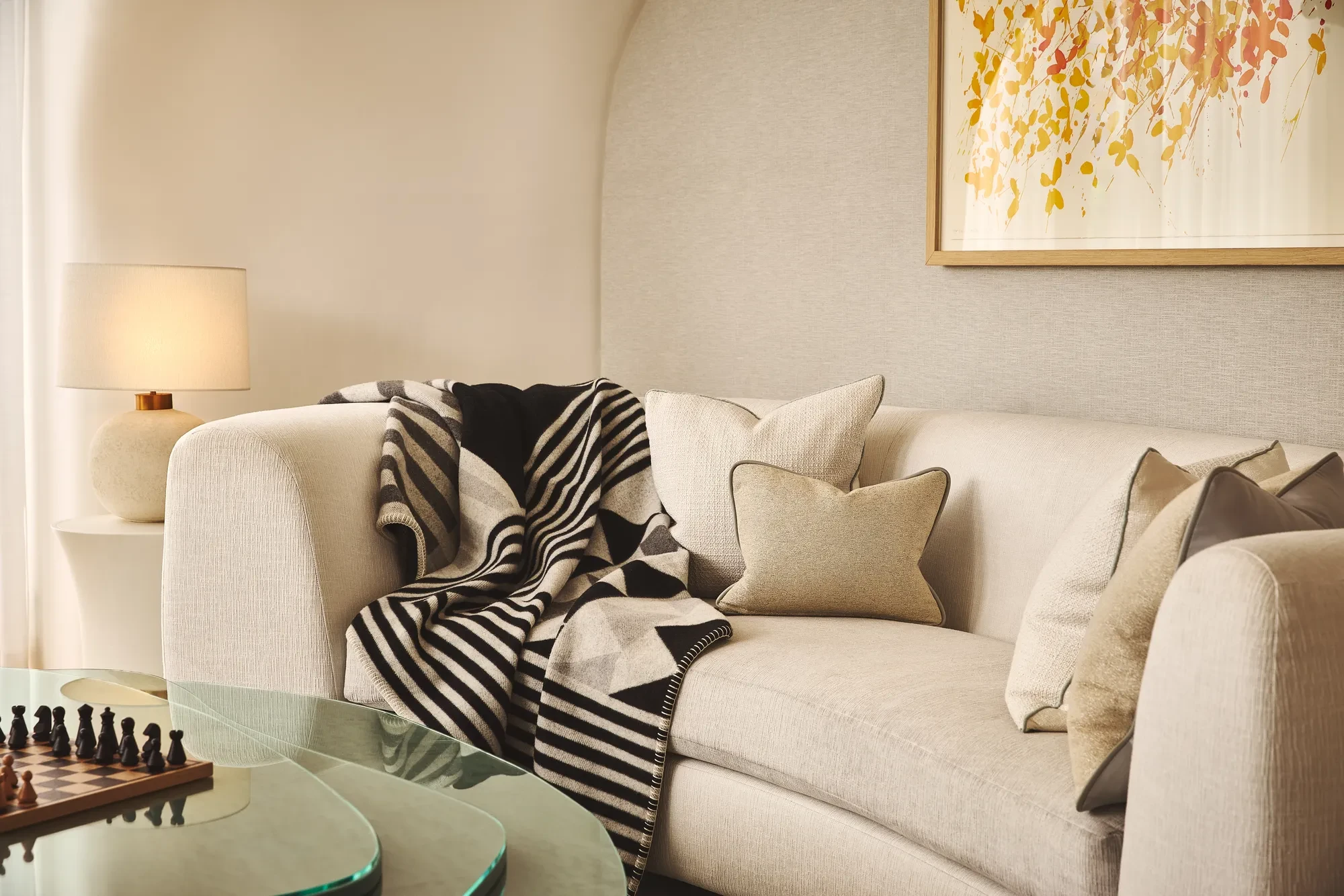 A cosy living area with a cream curved sofa layered with neutral cushions and a black-and-white geometric throw, beside a glass coffee table with a chessboard and a soft-glowing lamp on a side table.