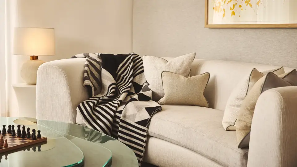 A cosy living area with a cream curved sofa layered with neutral cushions and a black-and-white geometric throw, beside a glass coffee table with a chessboard and a soft-glowing lamp on a side table.