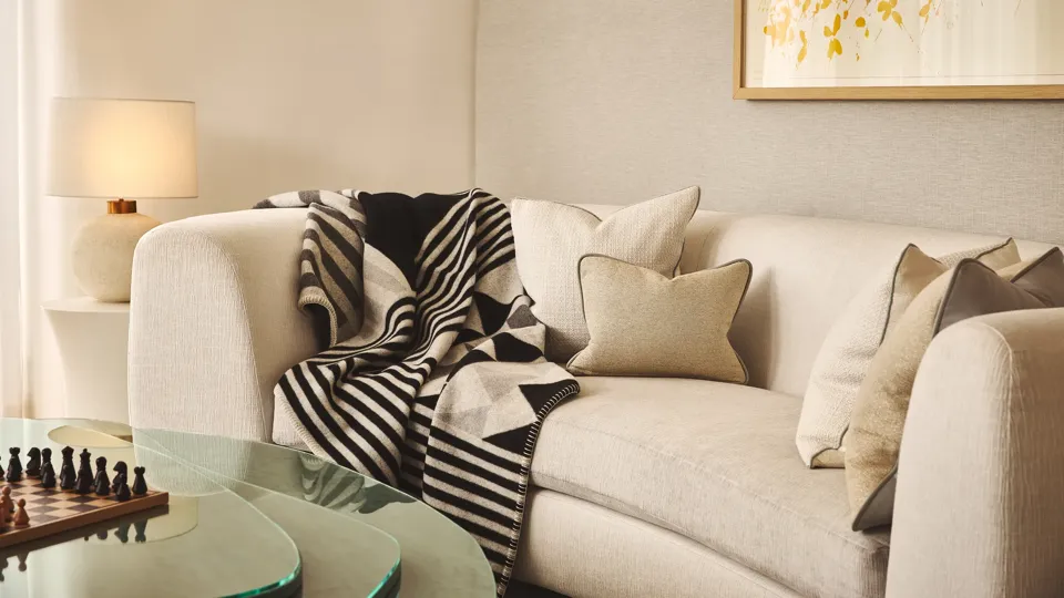 A cosy living area with a cream curved sofa layered with neutral cushions and a black-and-white geometric throw, beside a glass coffee table with a chessboard and a soft-glowing lamp on a side table.