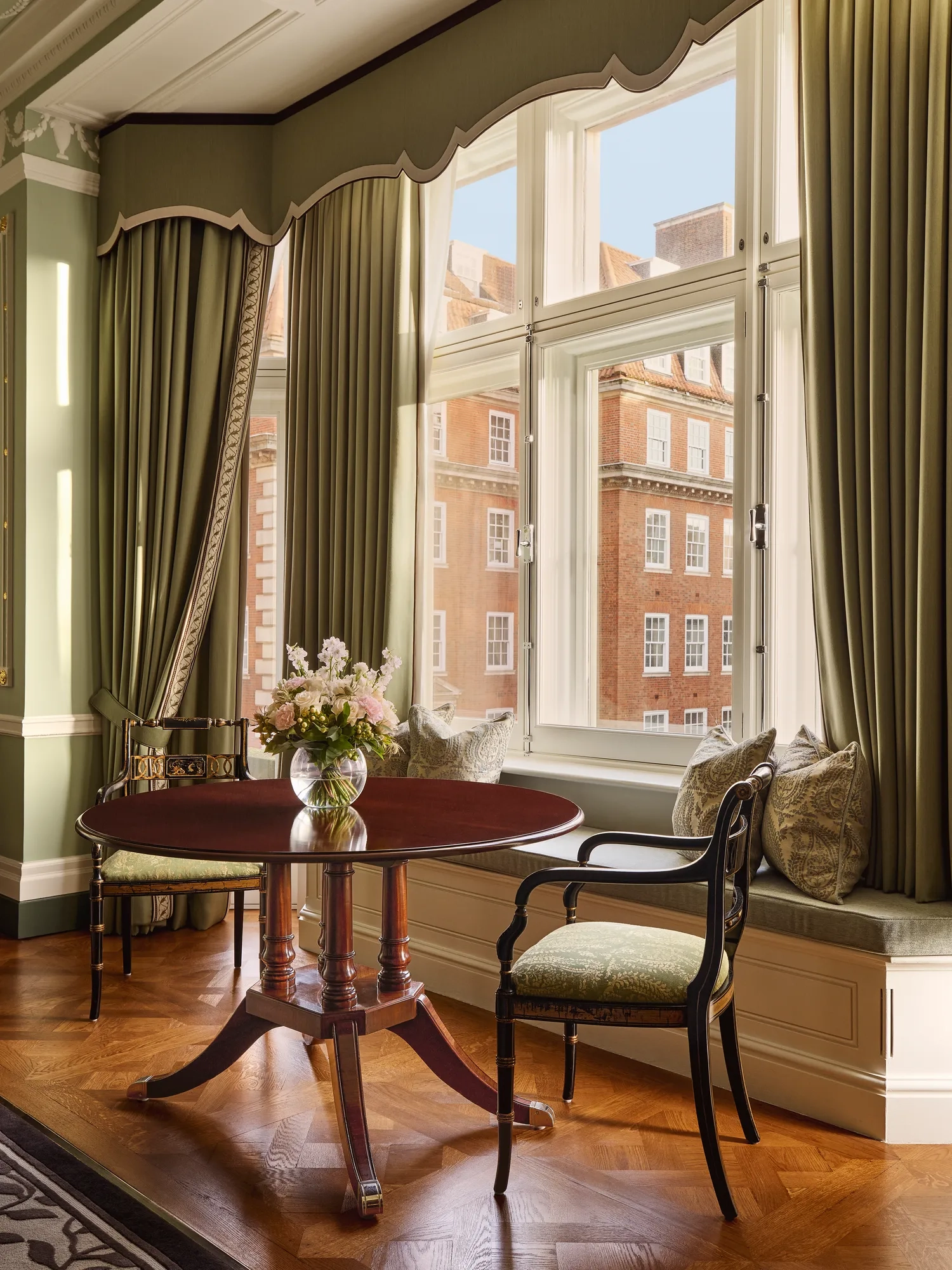 Elegant sitting area with round table and window seat overlooking red-brick buildings.