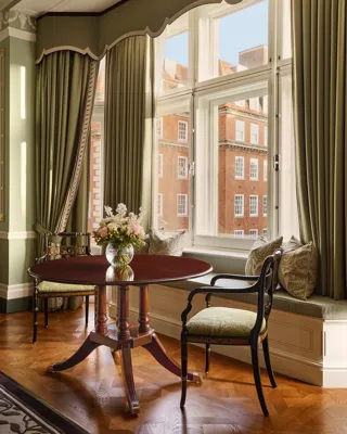 Elegant sitting area with round table and window seat overlooking red-brick buildings.