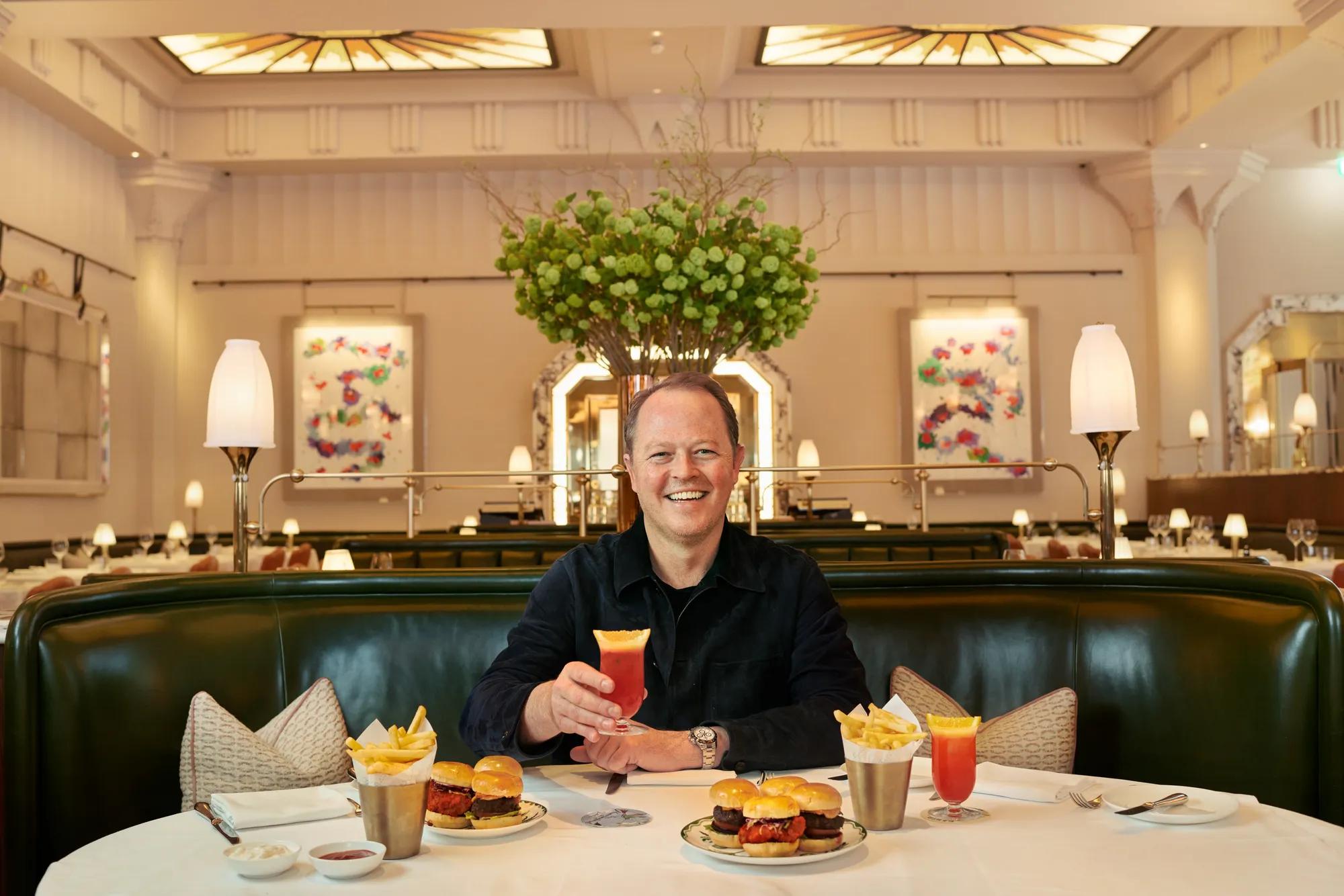 Man seated in elegant restaurant booth holding a cocktail, with sliders, fries and matching drinks on a white tablecloth