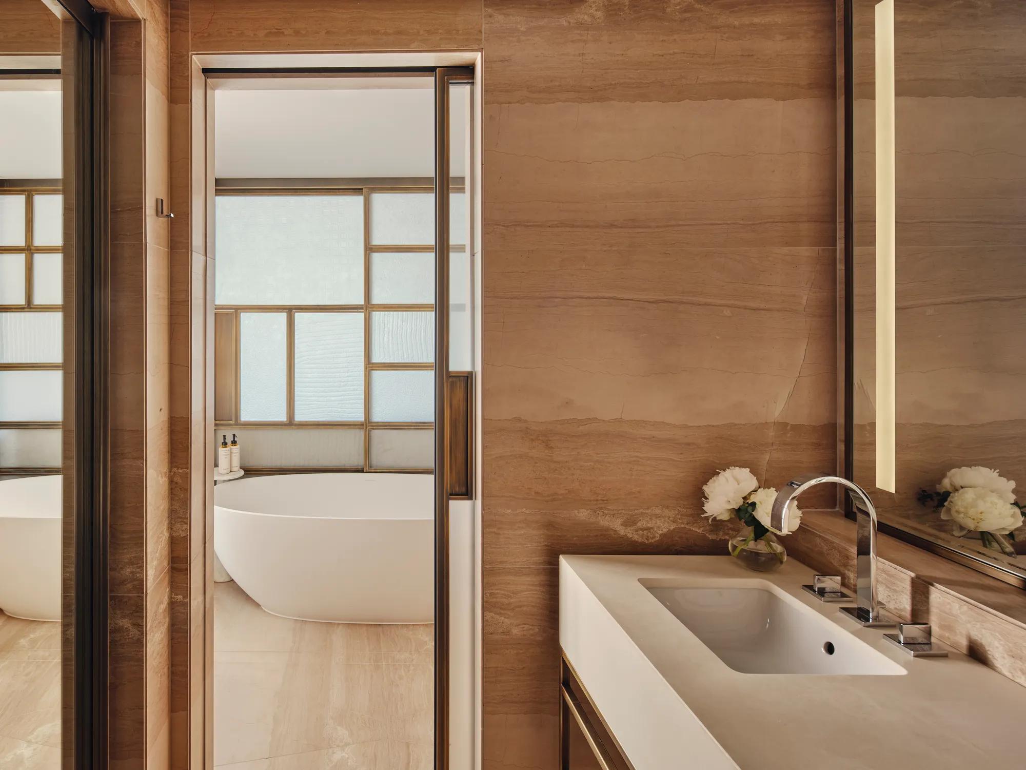 A marble bathroom with a sink and mirror, and a bath in the back