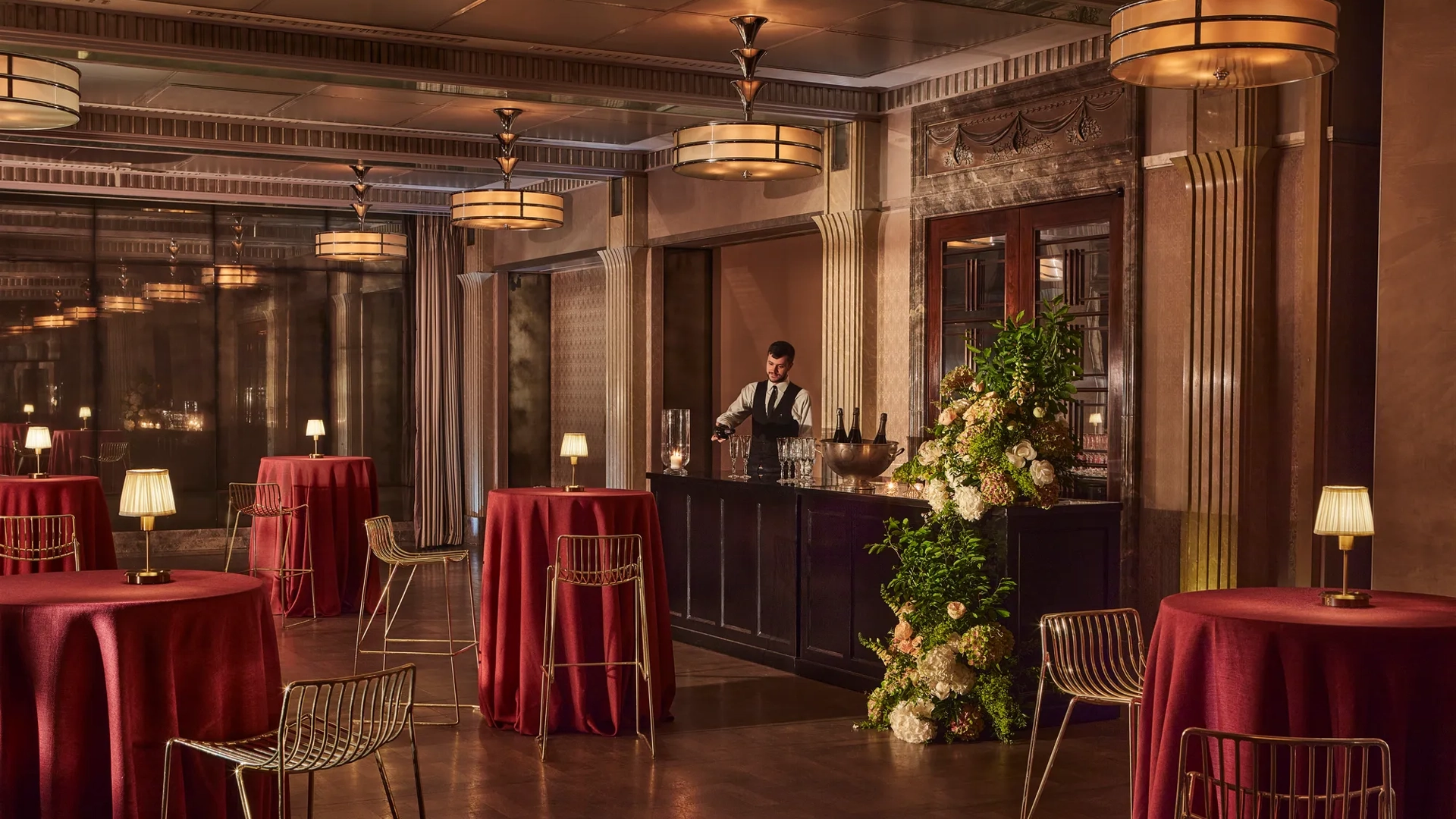 Elegant event bar with tall round tables draped in red cloth, gold chairs, and a bartender behind a floral-decorated bar.