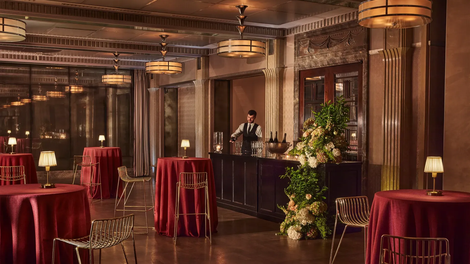 Elegant event bar with tall round tables draped in red cloth, gold chairs, and a bartender behind a floral-decorated bar.