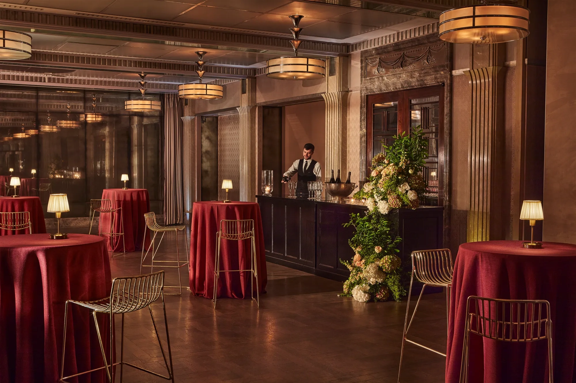 Elegant event bar with tall round tables draped in red cloth, gold chairs, and a bartender behind a floral-decorated bar.