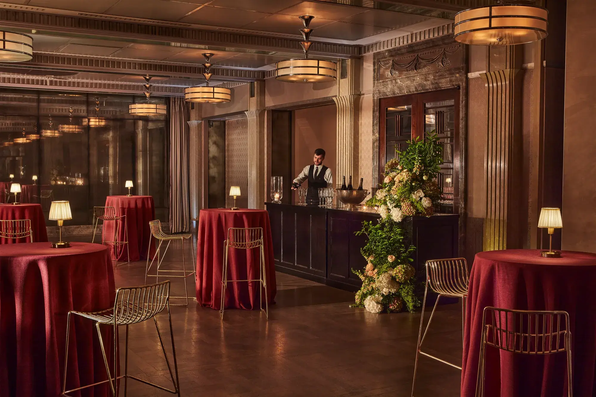 Elegant event bar with tall round tables draped in red cloth, gold chairs, and a bartender behind a floral-decorated bar.