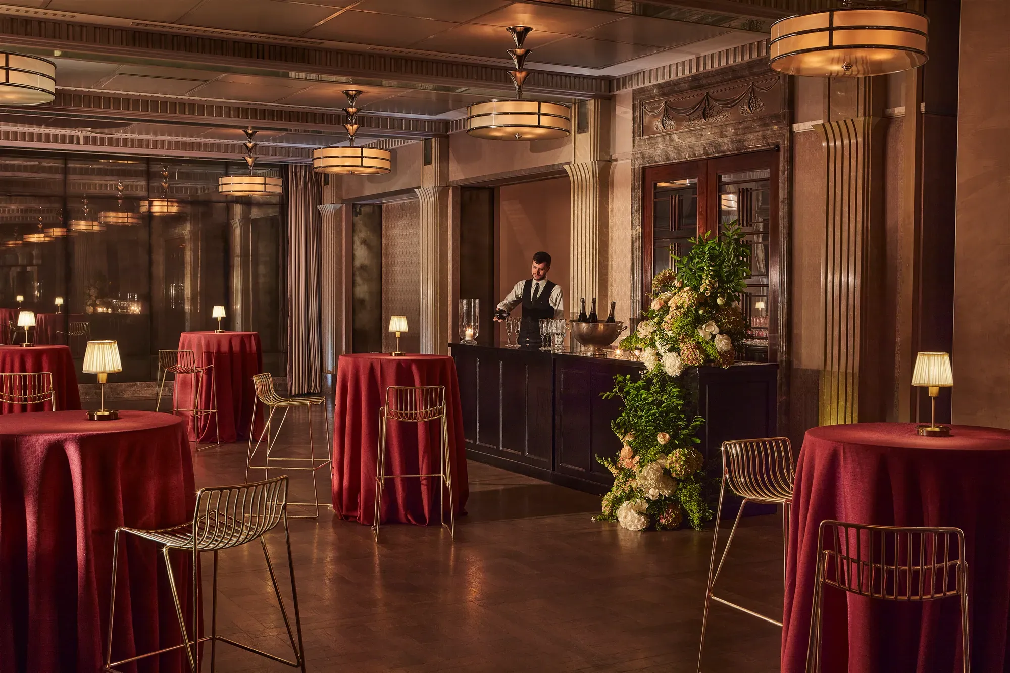 Elegant event bar with tall round tables draped in red cloth, gold chairs, and a bartender behind a floral-decorated bar.