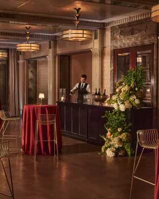 Elegant event bar with tall round tables draped in red cloth, gold chairs, and a bartender behind a floral-decorated bar.