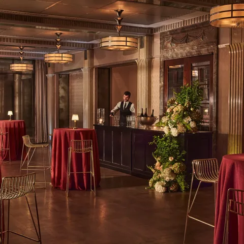Elegant event bar with tall round tables draped in red cloth, gold chairs, and a bartender behind a floral-decorated bar.