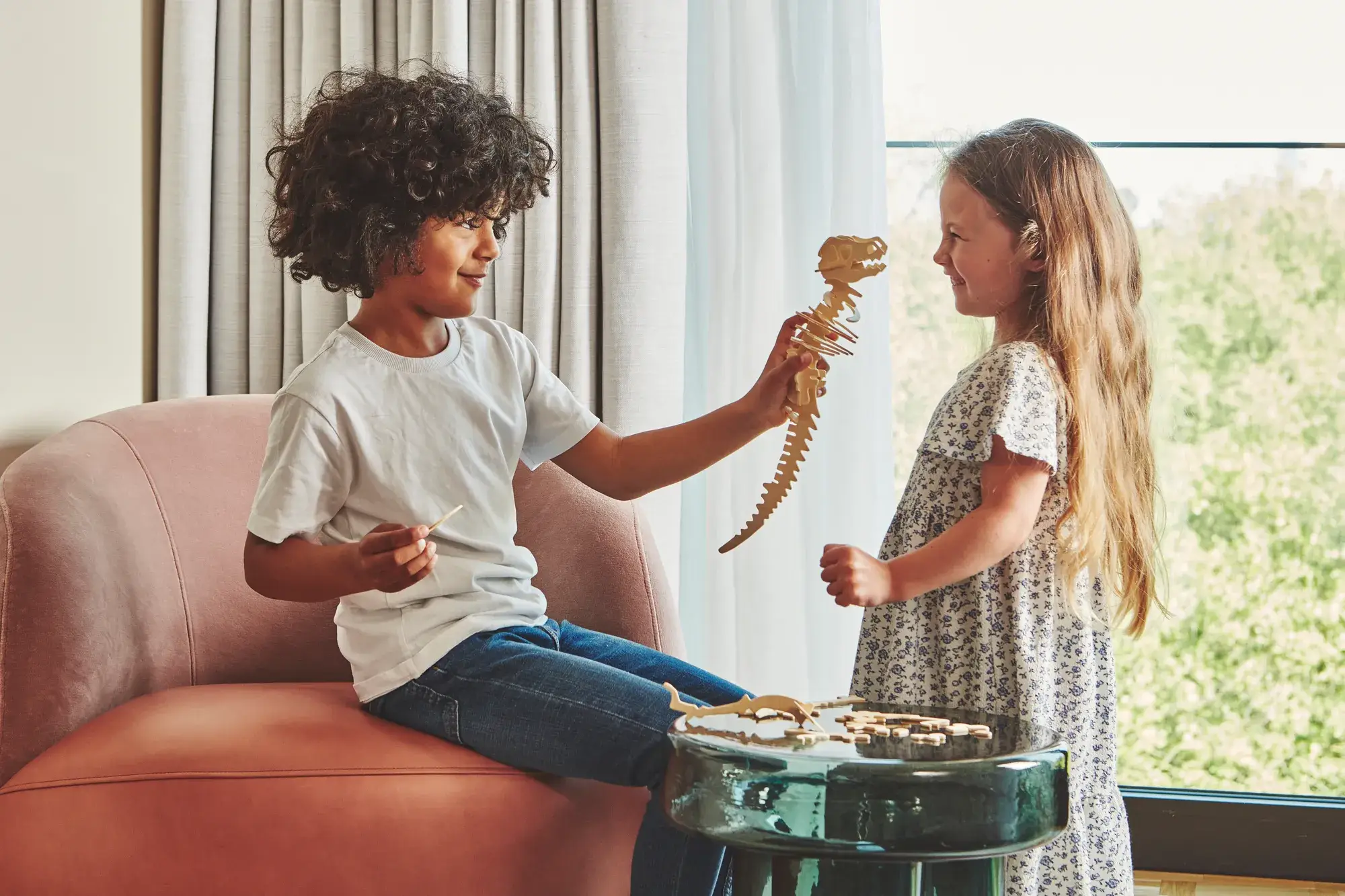 Two children playing with a wooden dinosaur puzzle by a window, seated on an armchair in a bright hotel room.