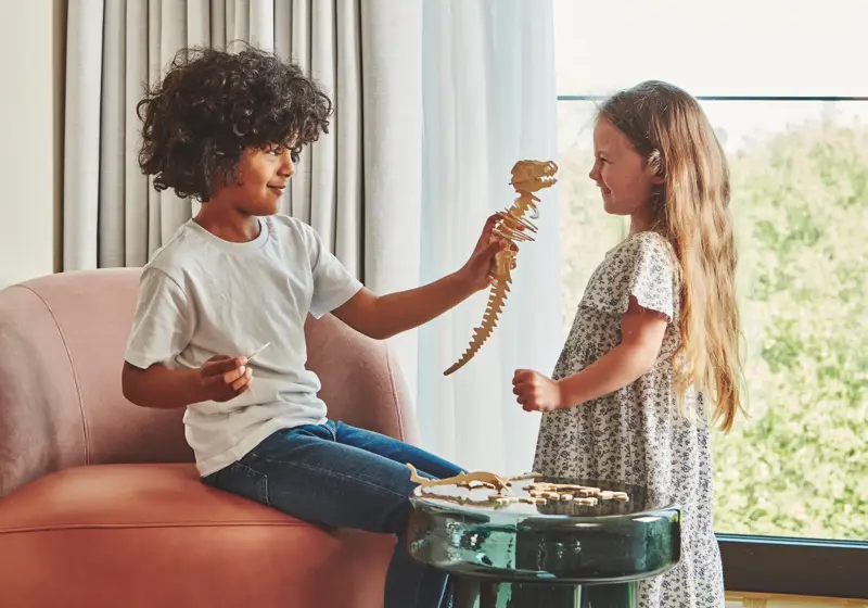 Two children playing with a wooden dinosaur puzzle by a window, seated on an armchair in a bright hotel room.