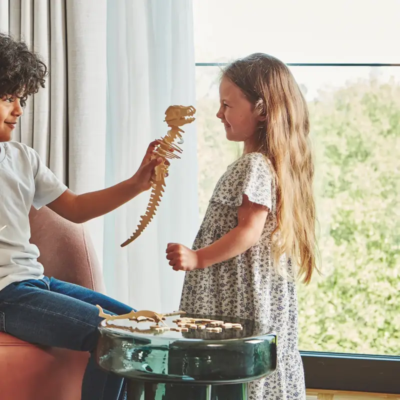Two children playing with a wooden dinosaur puzzle by a window, seated on an armchair in a bright hotel room.