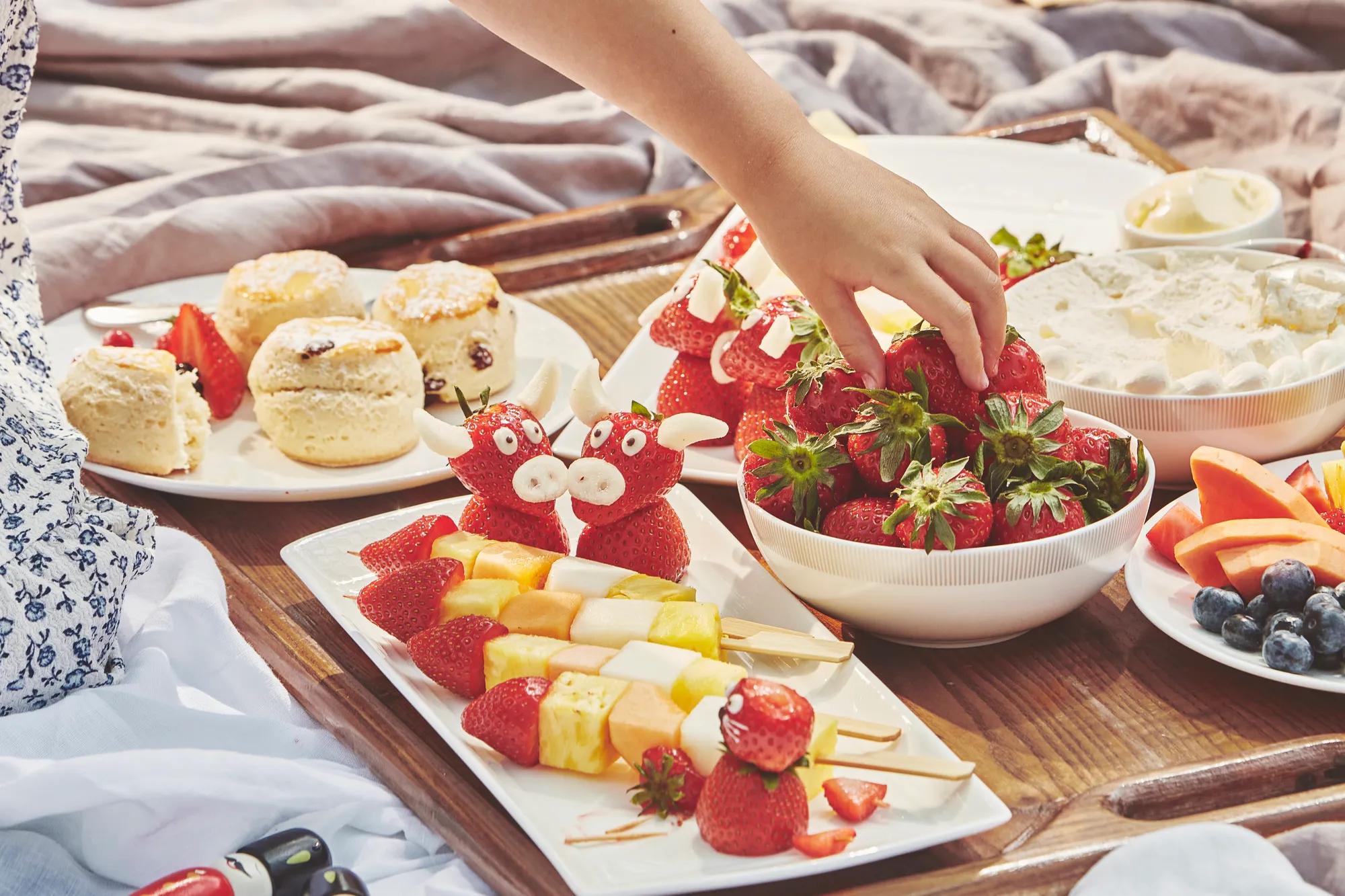 Child’s hand reaches for strawberries on a tray with fruit skewers, scones, and playful strawberry treats.
