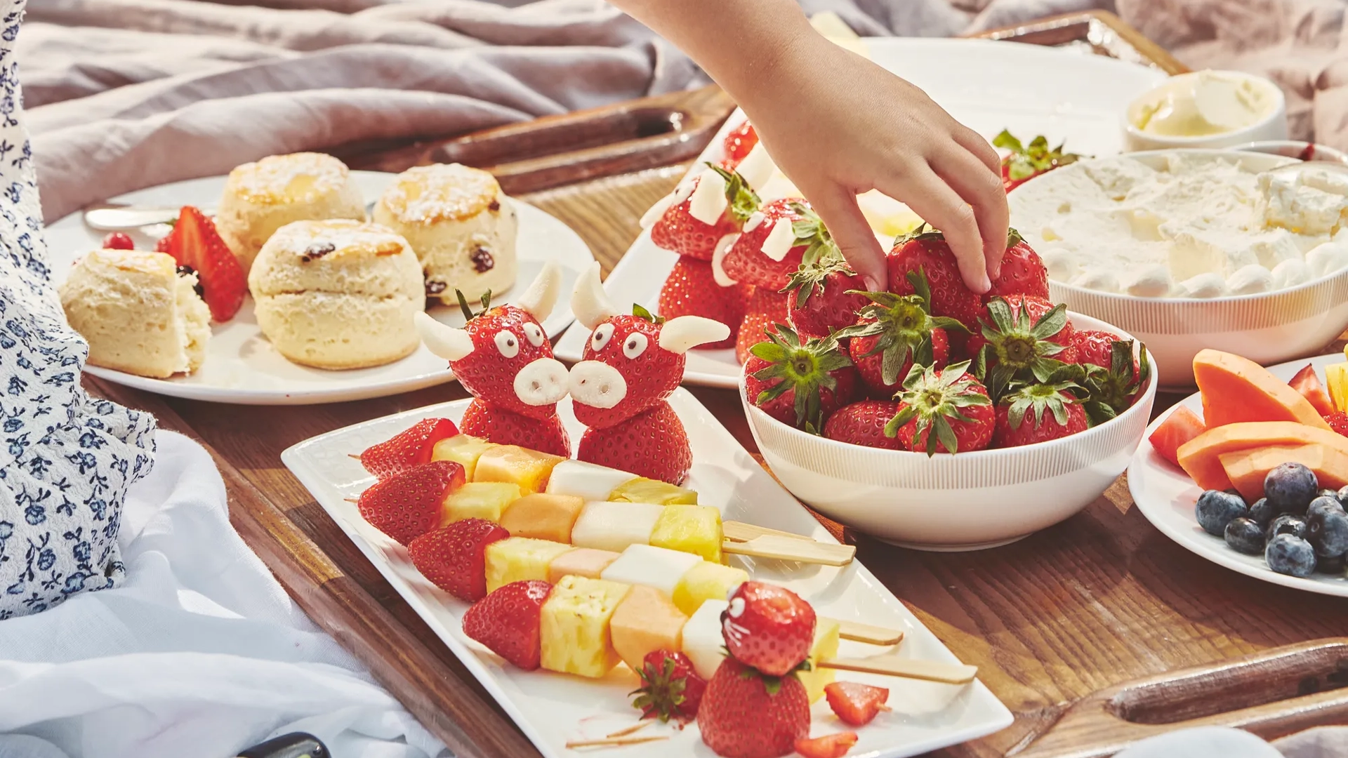 Child’s hand reaches for strawberries on a tray with fruit skewers, scones, and playful strawberry treats.