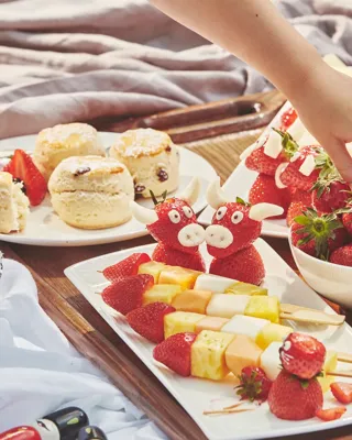 Child’s hand reaches for strawberries on a tray with fruit skewers, scones, and playful strawberry treats.