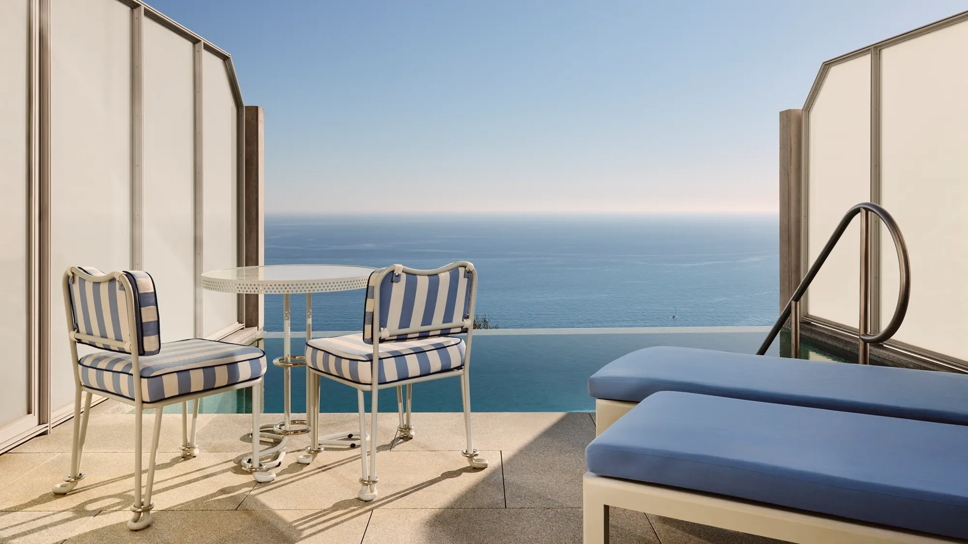 Private terrace with striped chairs, small round table, and sun loungers overlooking the calm blue sea.