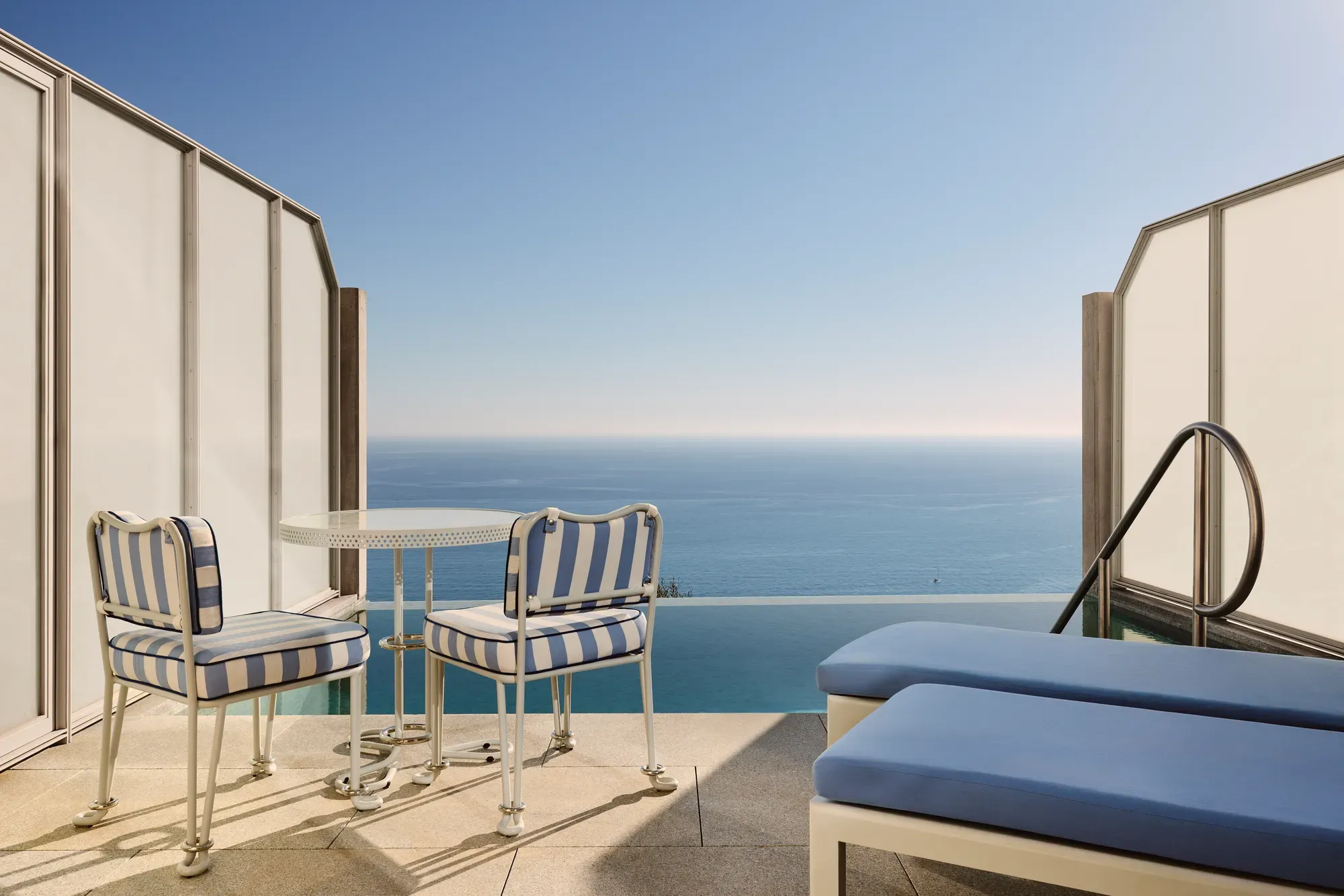 Private terrace with striped chairs, small round table, and sun loungers overlooking the calm blue sea.
