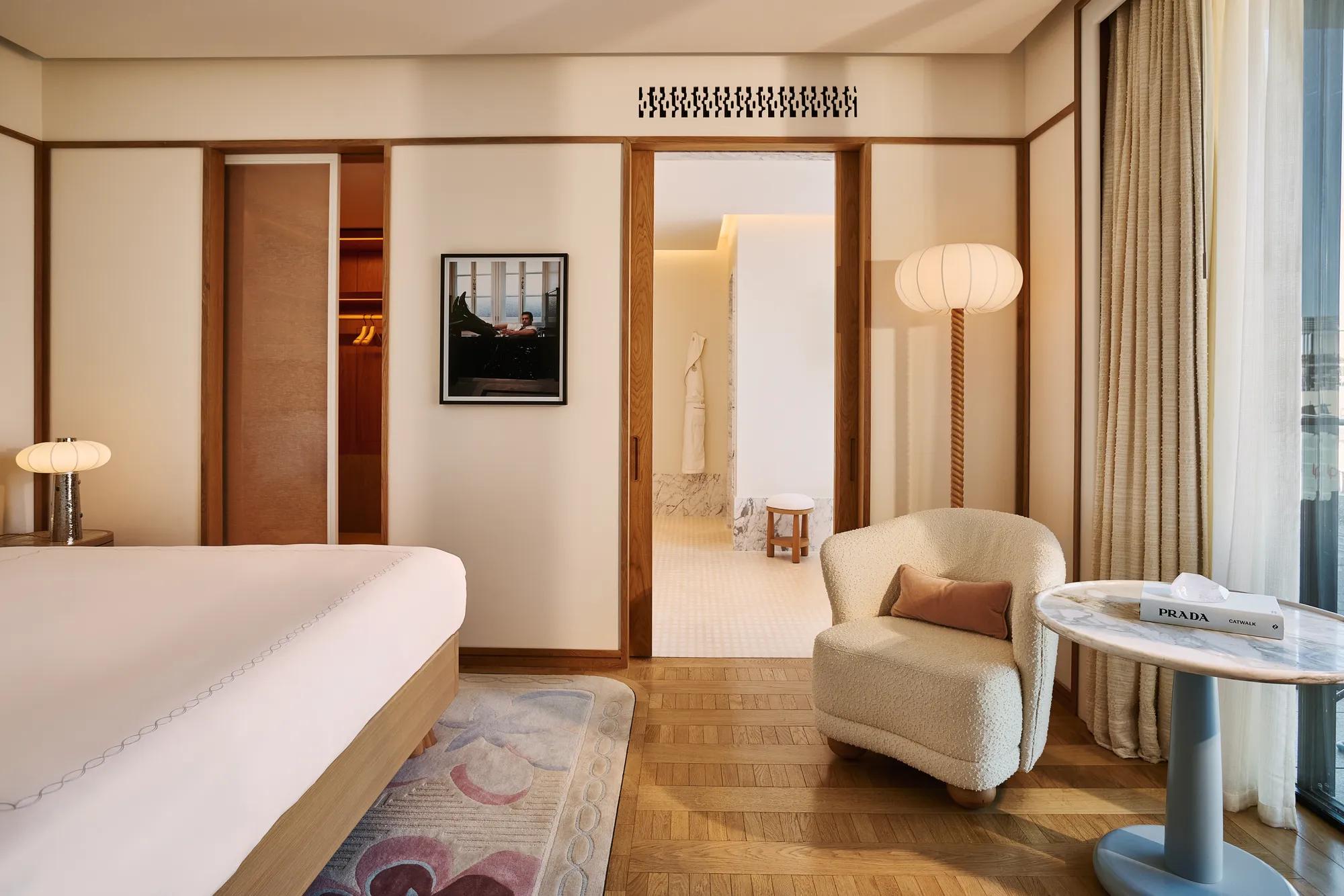 Serene bedroom with bed and armchair, wooden floors, sliding doors to bathroom, framed artwork, and side table by the window.