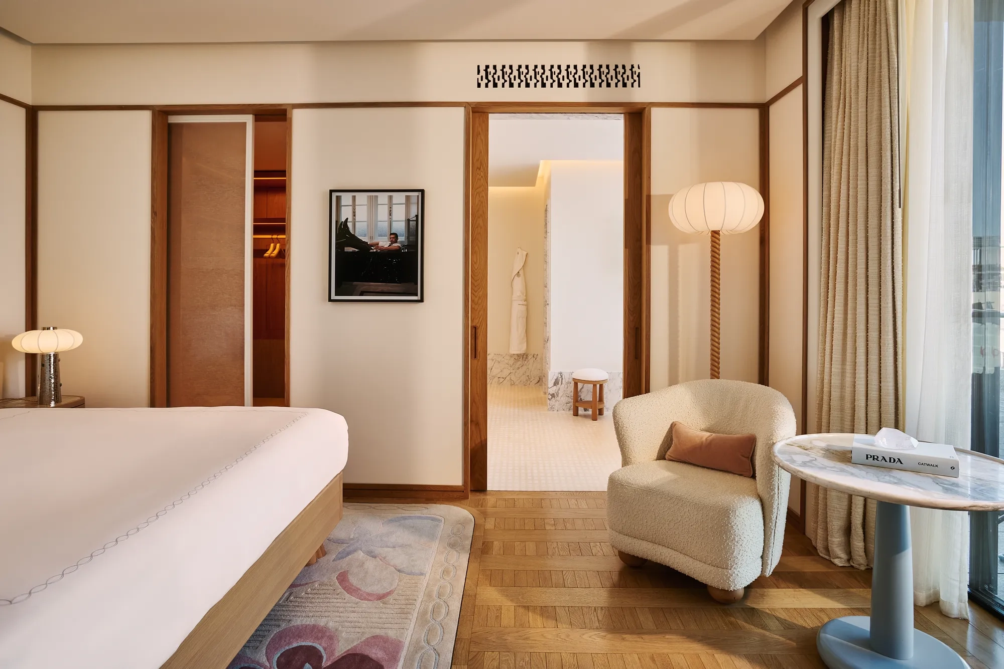 Serene bedroom with bed and armchair, wooden floors, sliding doors to bathroom, framed artwork, and side table by the window.
