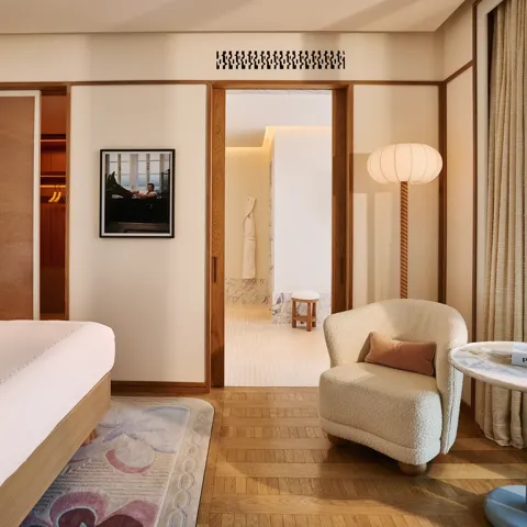 Serene bedroom with bed and armchair, wooden floors, sliding doors to bathroom, framed artwork, and side table by the window.