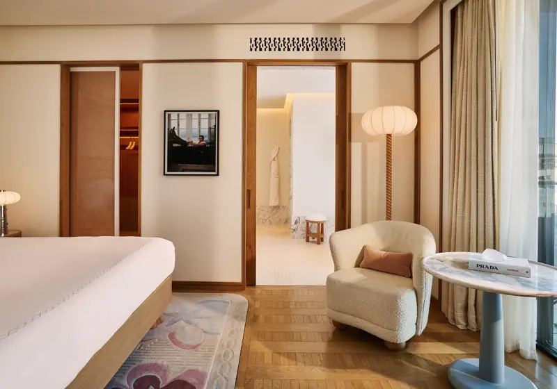 Serene bedroom with bed and armchair, wooden floors, sliding doors to bathroom, framed artwork, and side table by the window.