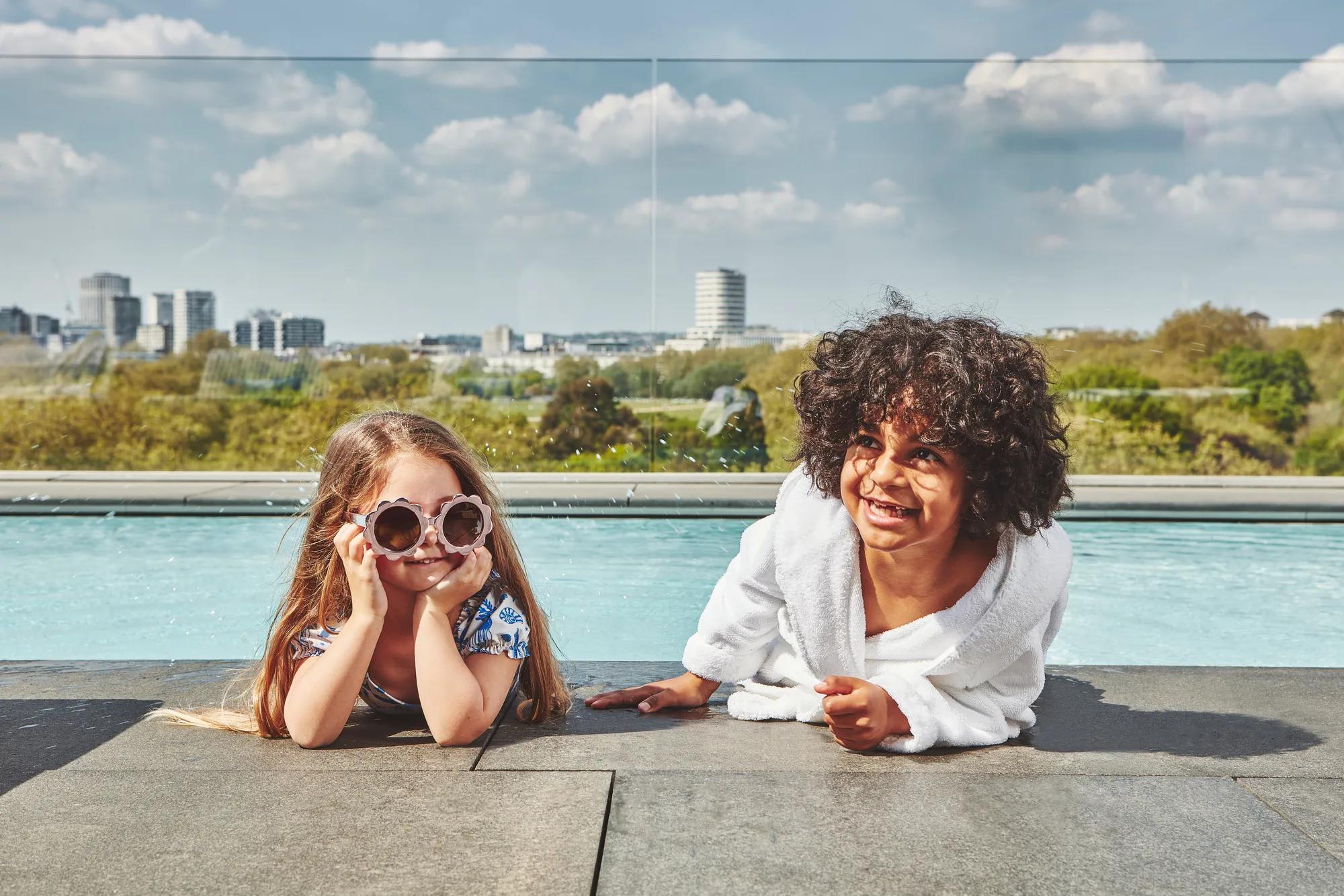 Two children relaxing by a rooftop pool with city skyline in the background.