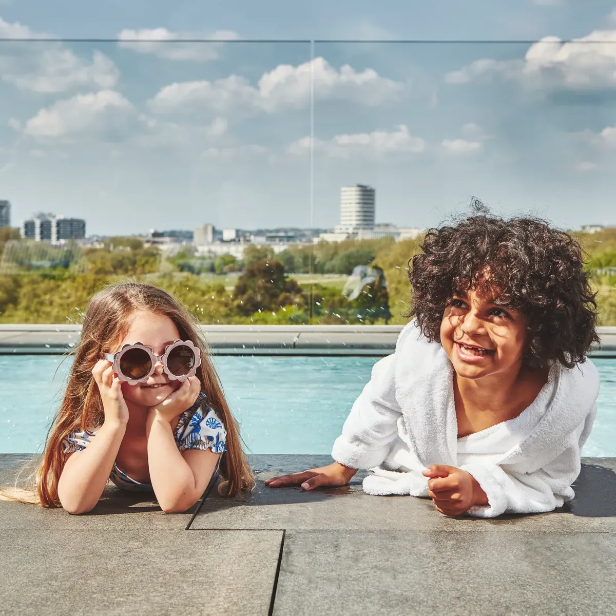 Two children relaxing by a rooftop pool with city skyline in the background.