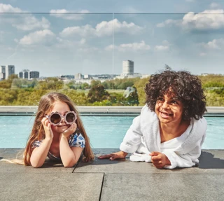 Two children relaxing by a rooftop pool with city skyline in the background.