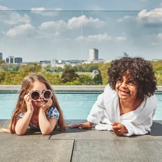 Two children relaxing by a rooftop pool with city skyline in the background.