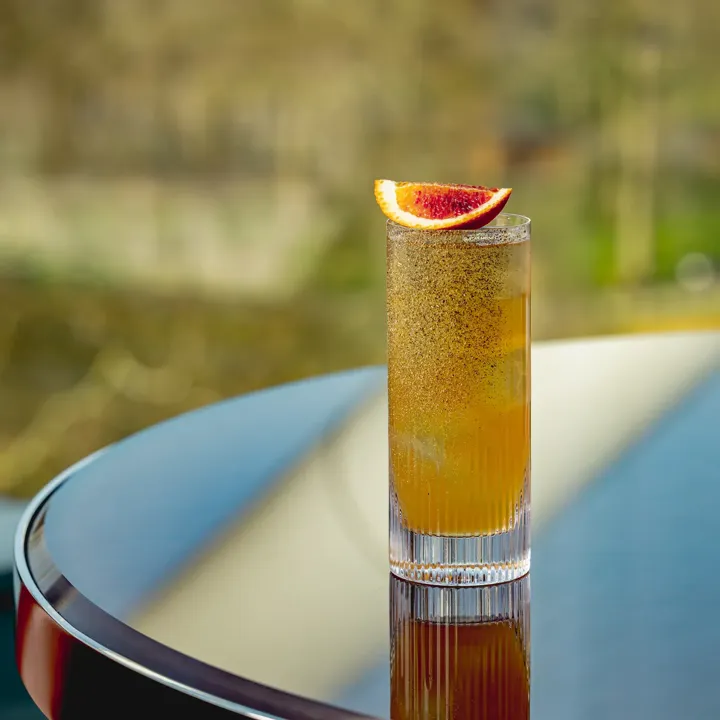 Golden cocktail garnished with blood orange slice on a reflective table.